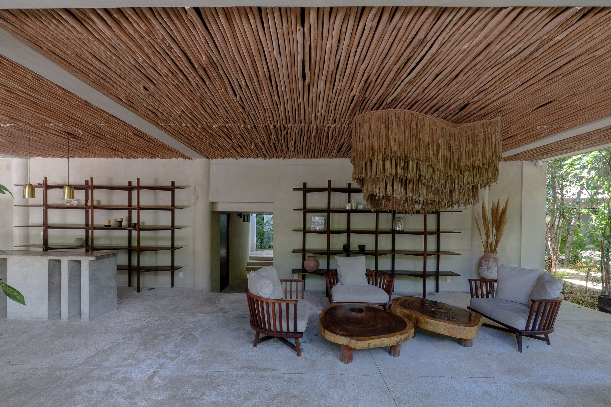 Open-air common lounge with a woven ceiling, shelving, armchairs and round wooden tables