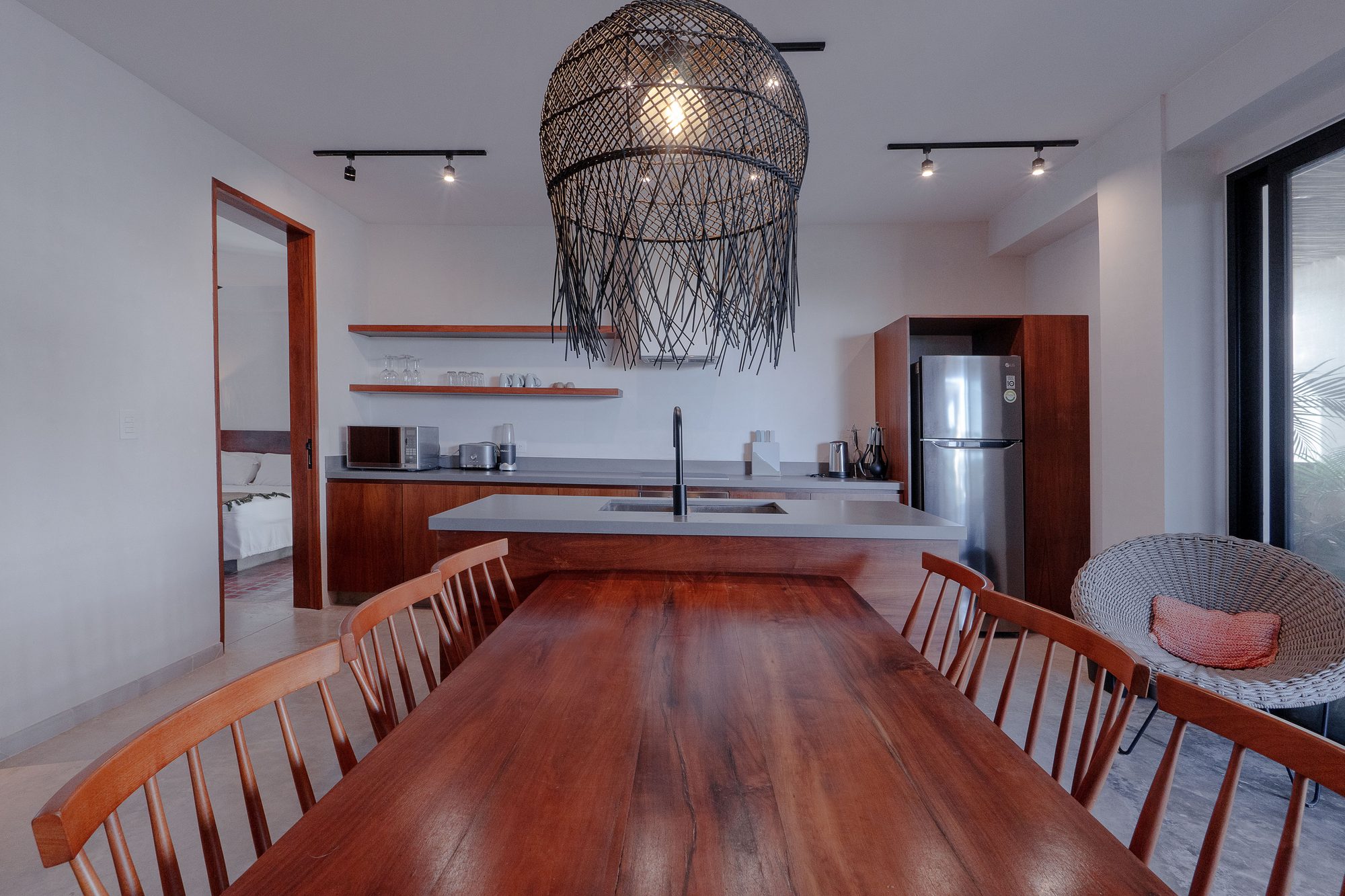 Wooden dining table under a woven pendant lamp, with the kitchen behind