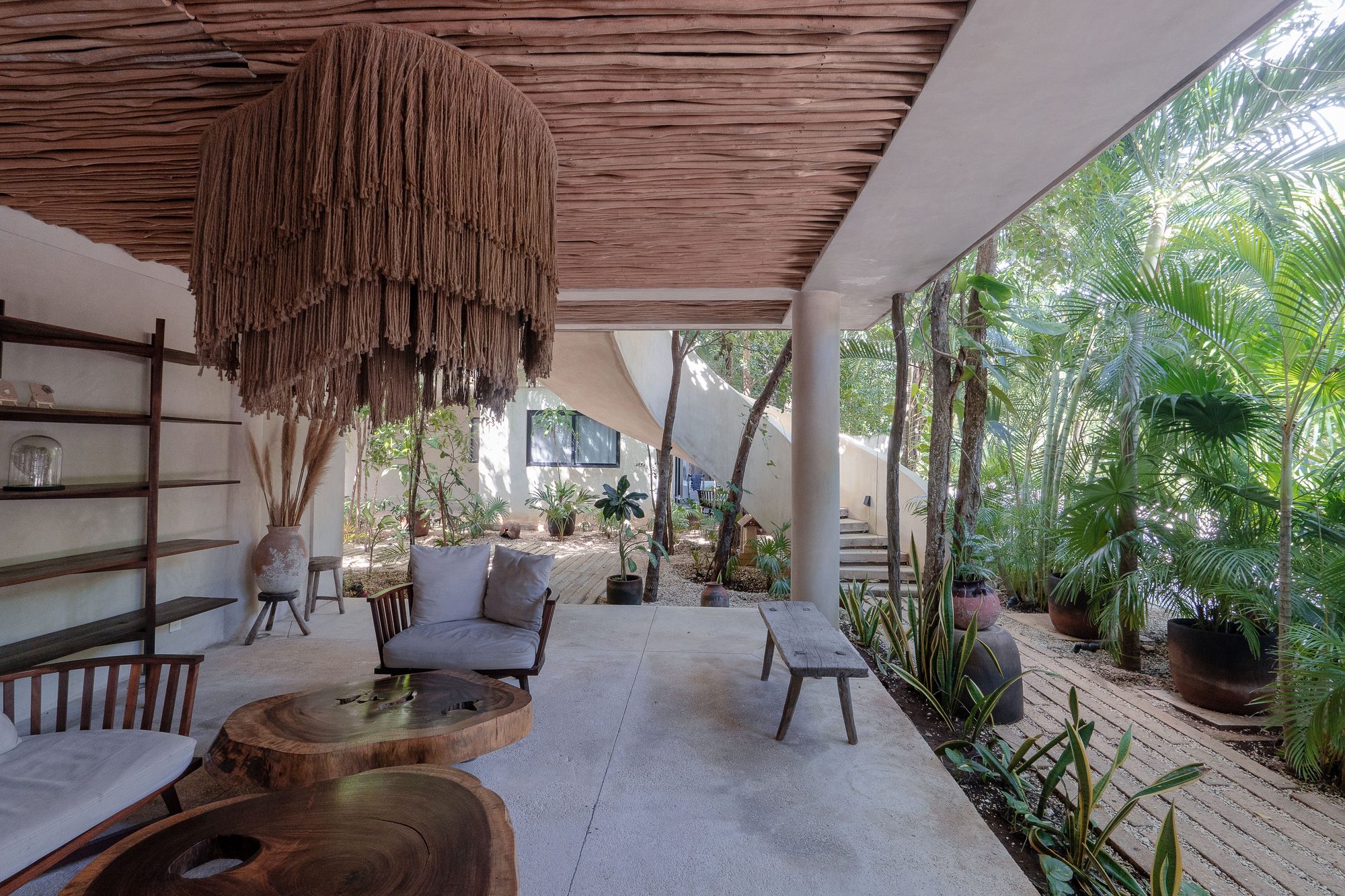 Shaded lounge with a large woven pendant, wood slab tables and armchairs beside a garden path